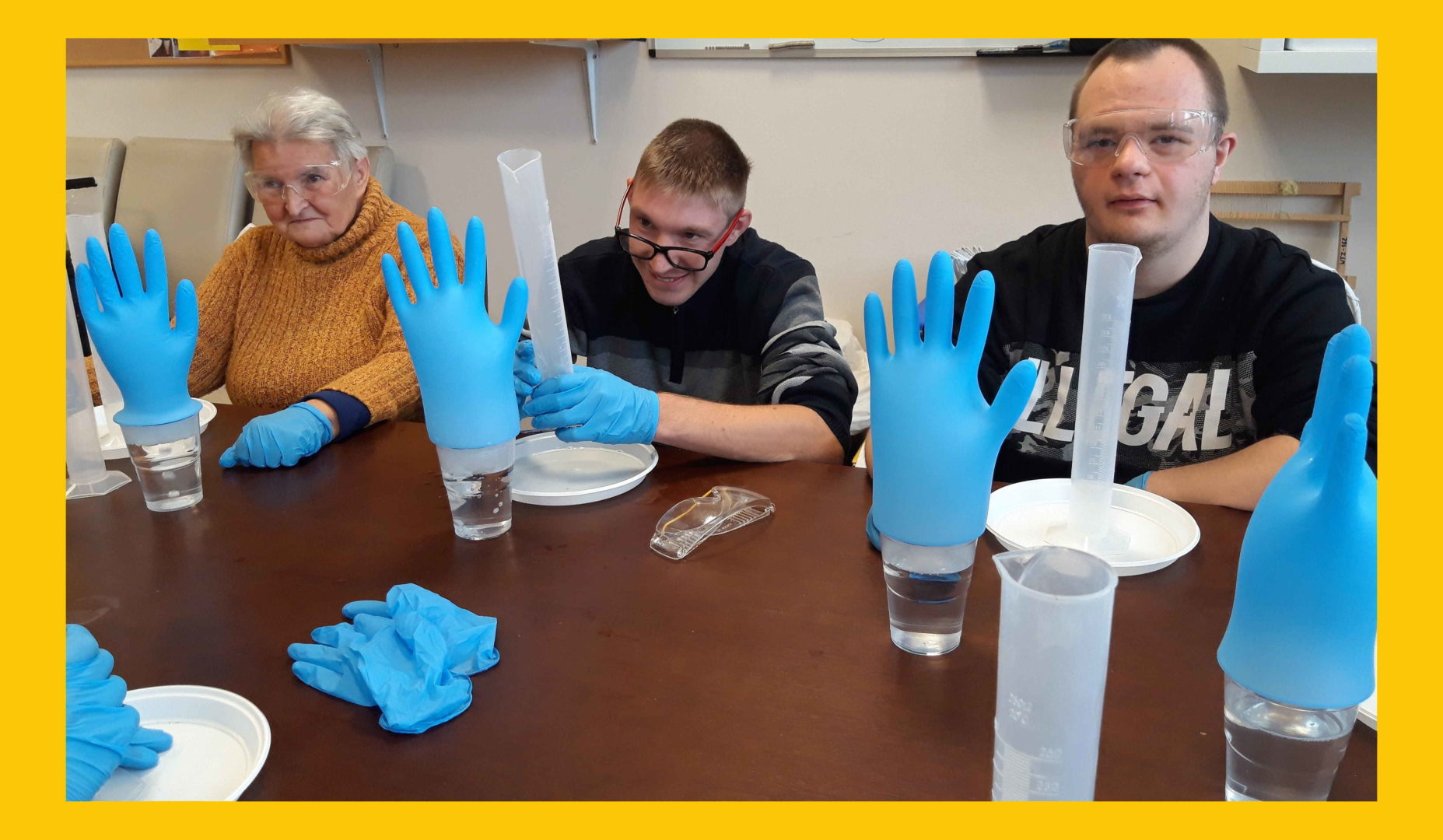 Na zdjęciu trzy osoby siedzące za stołem, robiące eksperyment. Na szklanki nałożona jest gumowa rękawiczka.