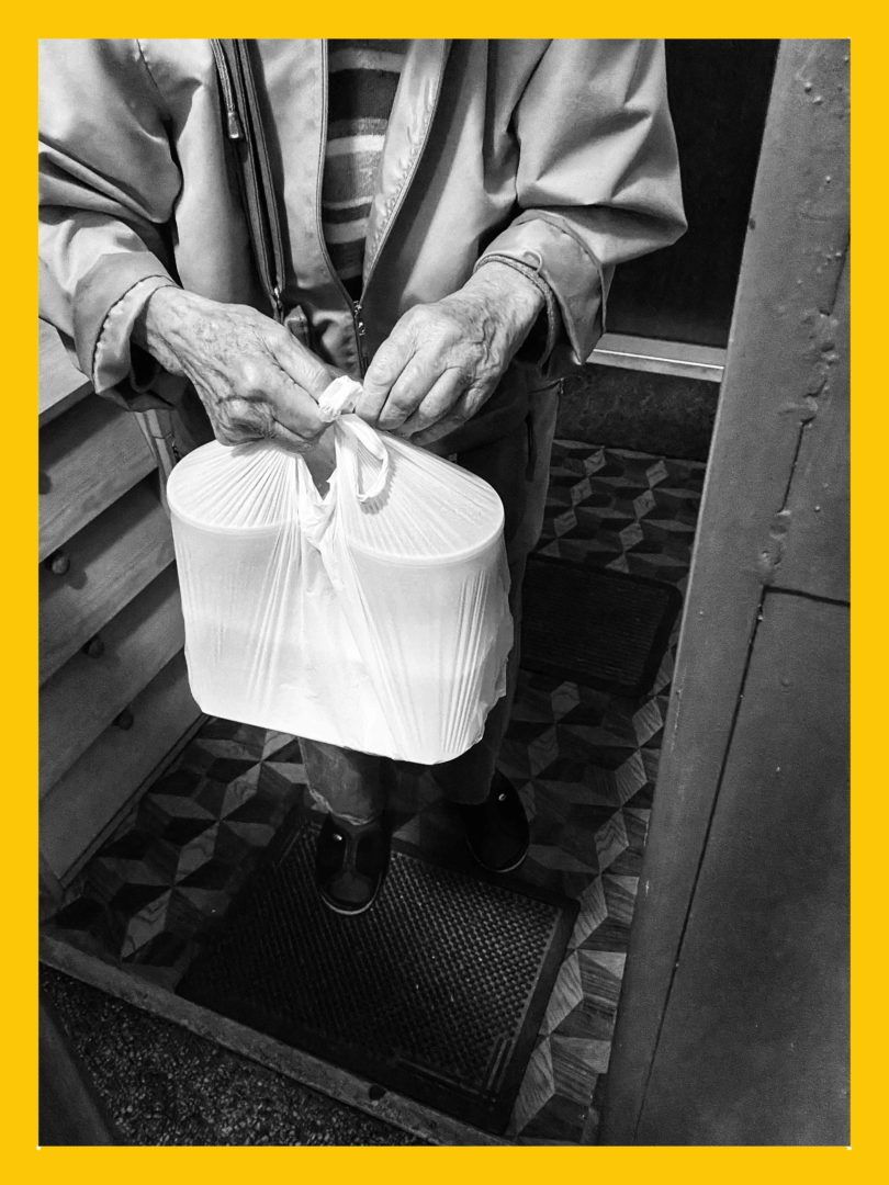 Zdjęcie przedstawia dłonie starszej osoby, trzymające siatkę z pojemnikami z żywnością w progu mieszkania.