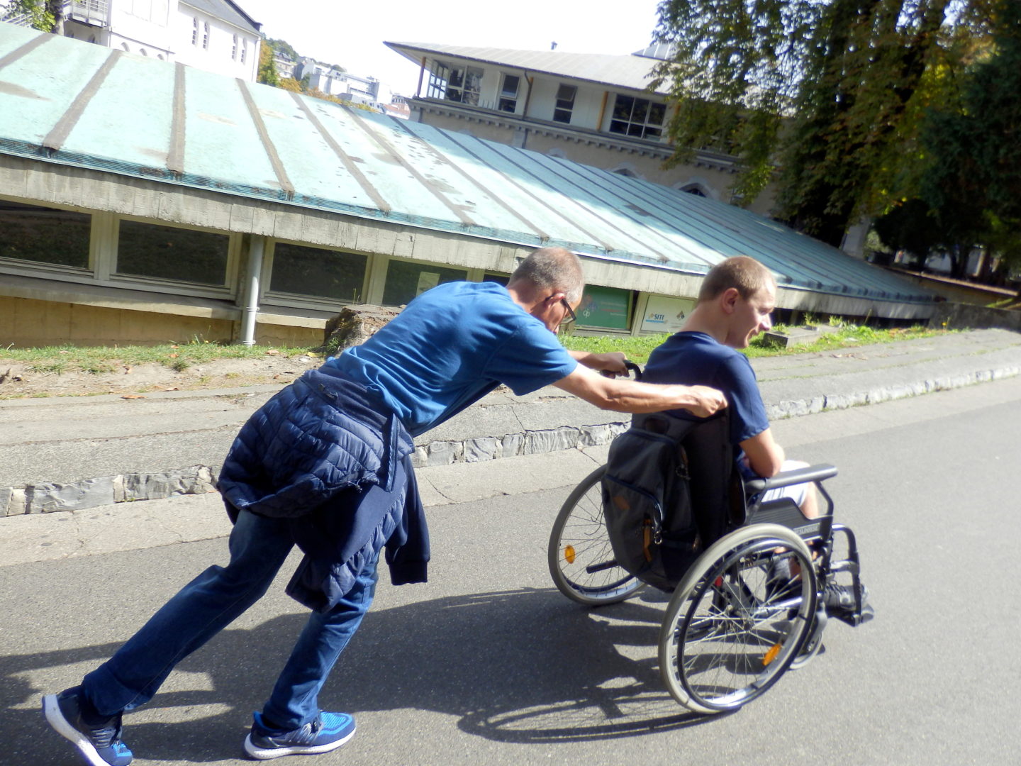 Na zdjęciu mężczyzna pchający wózek inwalidzki w drugim mężczyzną. Pcha wózek pod górę, widać po nim wysiłek fizyczny. W tle rozległy, płaski dach oraz budynek.