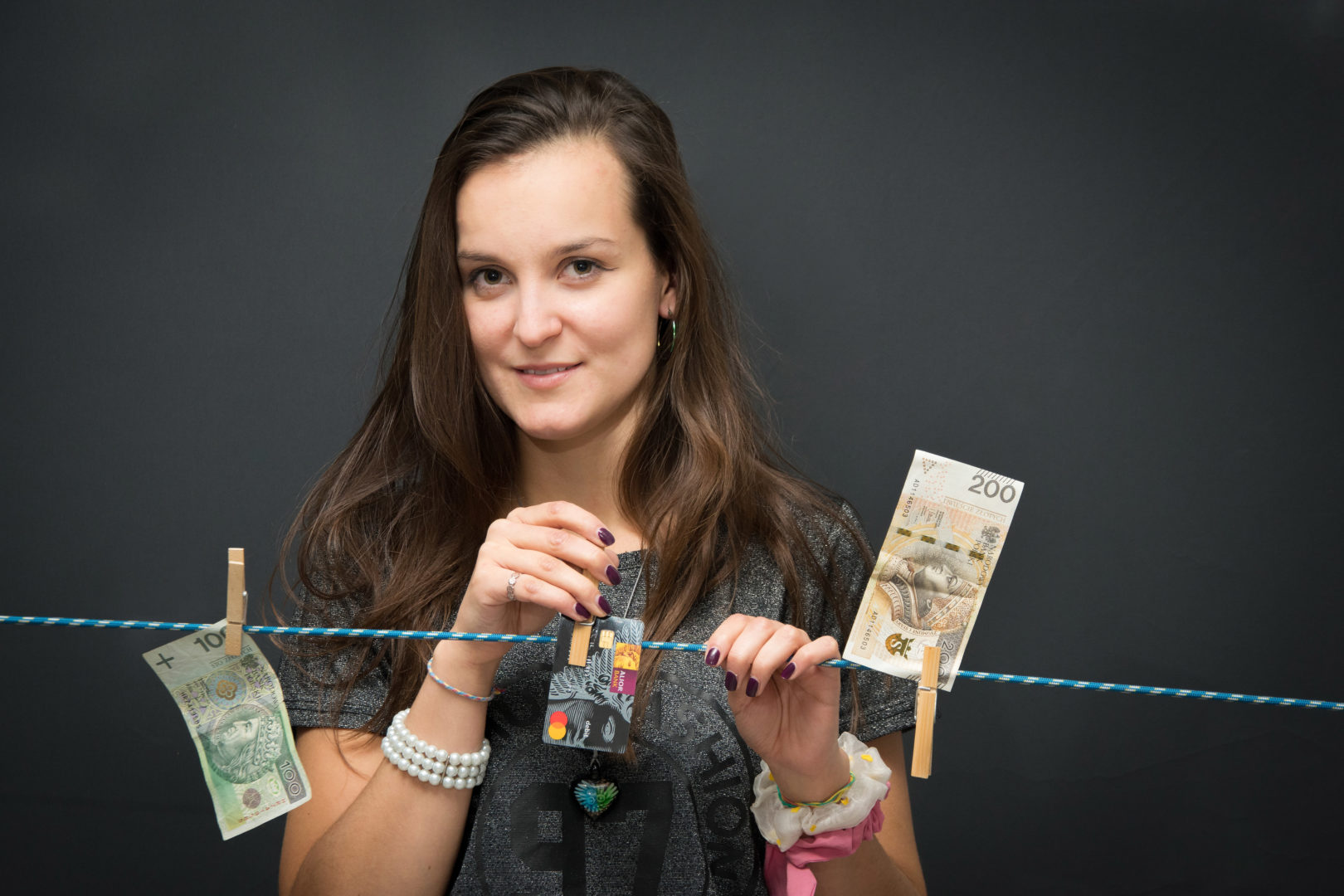 Na zdjęciu kobieta przypinająca na sznurku dwa banknoty w nominale 100 i 200 złotych. Kobieta widoczna jest od klatki piersiowej w górę, z tyłu ciemne tło.