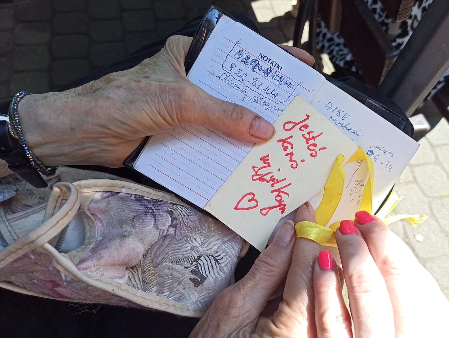 Na zdjęciu jest rozłożony mały kalendarz książkowy. Jest on trzymany w rękach starszej kobiety. Jej prawej dłoni dotyka dłoń młodej koboety, która jest wolontariuszem i pomaga starszej kobiecie. Na środku kalnedarza przyklejona jest karteczka z napisem "Jesteś kimś wyjątkowym" wraz z dorysowanym symbolem serca.