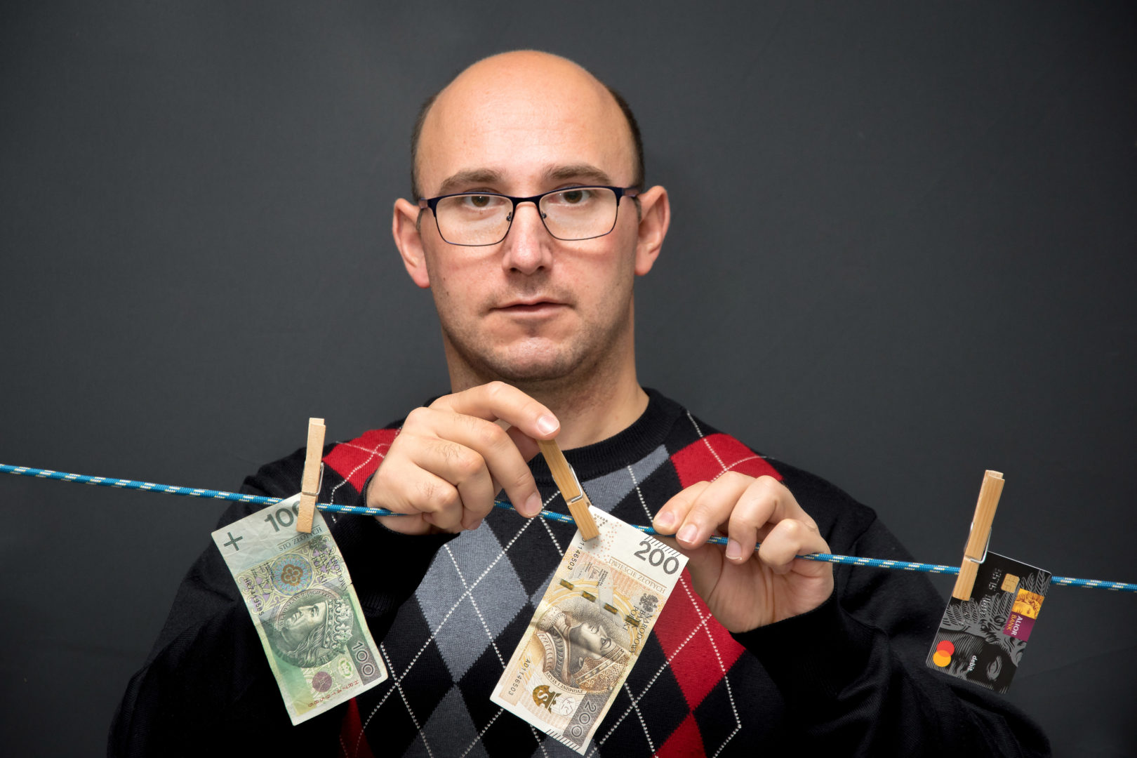 Na zdjęciu mężczyzna przypinający na sznurku dwa banknoty w nominale 100 i 200 złotych. Mężczyzna widoczny jest od torsu w górę, z tyłu ciemne tło.
