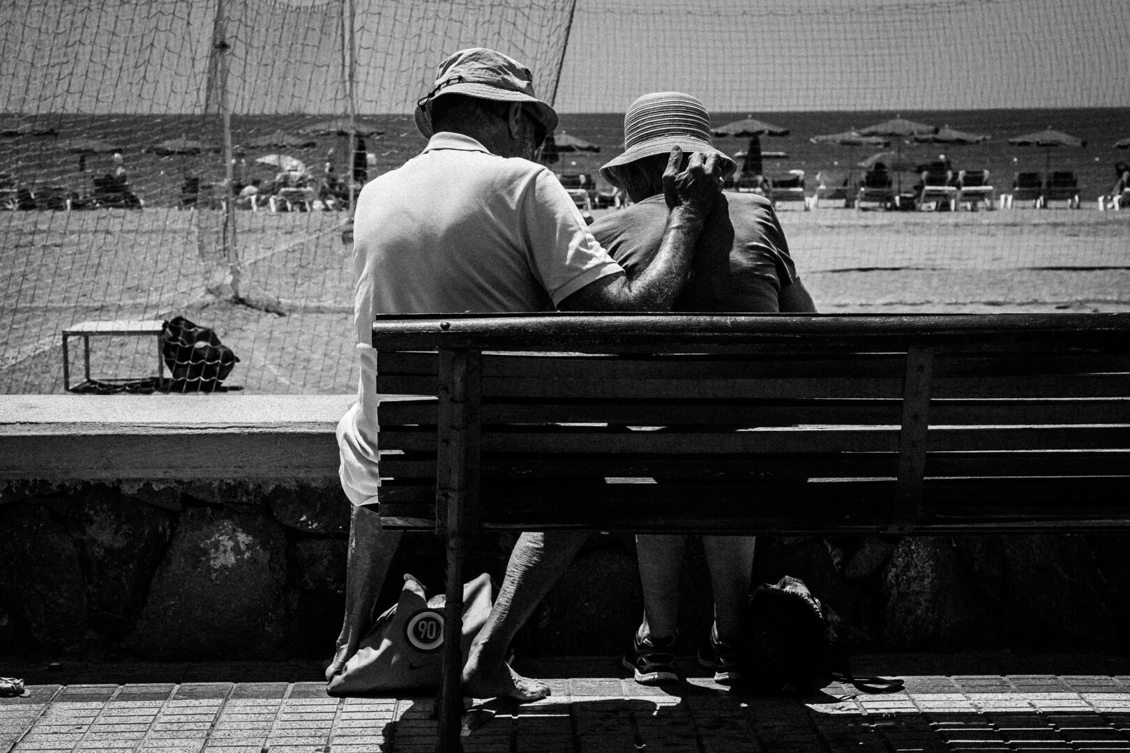 Na pierwszym planie lewa połowa ławki, ukazana od tyłu. Na lewym skraju siedzi starszy mężczyzna i trzyma przedramię na plecach kobiety siedzącej po prawej. Oboje mają letnie kapelusze, ubrani w bluzki z krótkim rękawem. W tle plaża, słomkowe parasole, leżaki i morze. Zdjęcie wykonane w technice czarno-białego zdjęcia.